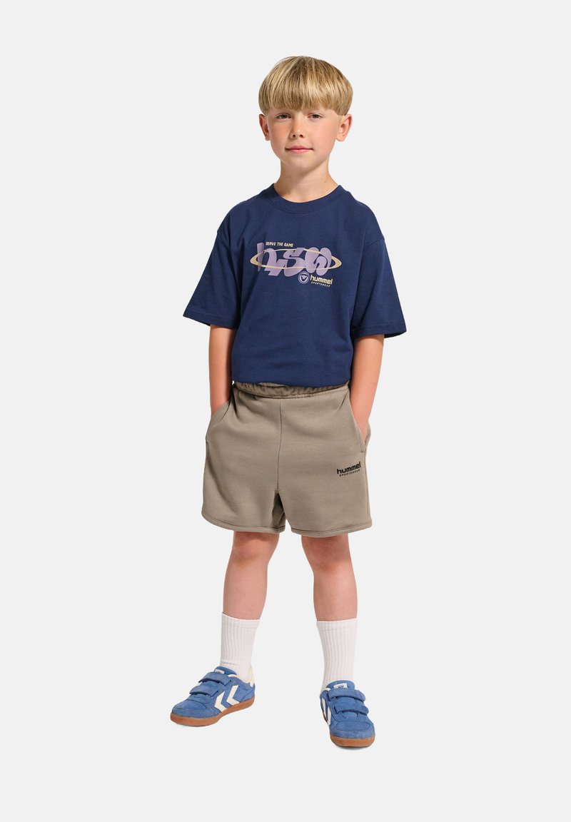 Marinfärgad grafisk t-shirt med lila tryck, beiga shorts, blå sneakers med vita detaljer och vita strumpor. Barnet står med händerna i shortsens fickor.