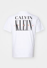 Valge puuvillane T-särk, mille seljal on suur must tekst "CALVIN KLEIN JEANS", kus sõna "JEANS" on esile tõstetud oranžina.