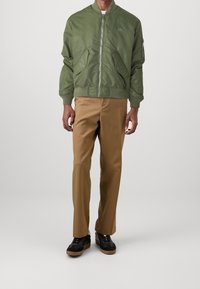 Zelená bomber bunda se zipem vpředu, žebrovanými manžetami, dvěma bočními kapsami a malým logem. Kombinována s hnědými kalhotami a černými teniskami.