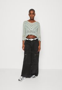 Mintgrün gehäkeltes Crop-Top mit ¾-Ärmeln, kombiniert mit schwarzen, weiten Cargo-Hosen mit Reißverschlüssen und Taschen, getragen mit mehrfarbigen Sneakers.