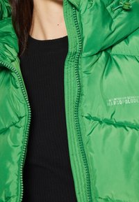 Zielona pikowana kurtka o błyszczącej fakturze, z przednim zamkiem błyskawicznym i sznurkami przy kapturze. Noszona na czarnej prążkowanej bluzce.
