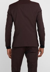 Donker bordeauxkleurig colbert, aansluitend ontwerp, textiel met structuur, met zwarte accentdetails op de polsen en knoopdetails op de mouwen. Achteraanzicht weergegeven.