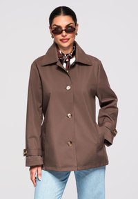 Kvinde iført brun jakke med knapper, lyseblå jeans, tændt halstørklæde med print og mørke solbriller, stående med hånden i lommen.