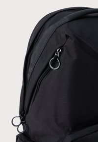 Mochila negra con un tejido texturizado, un bolsillo con cremallera y un tirador en forma de lazo, y bordes curvados. Diseño robusto con costuras reforzadas.