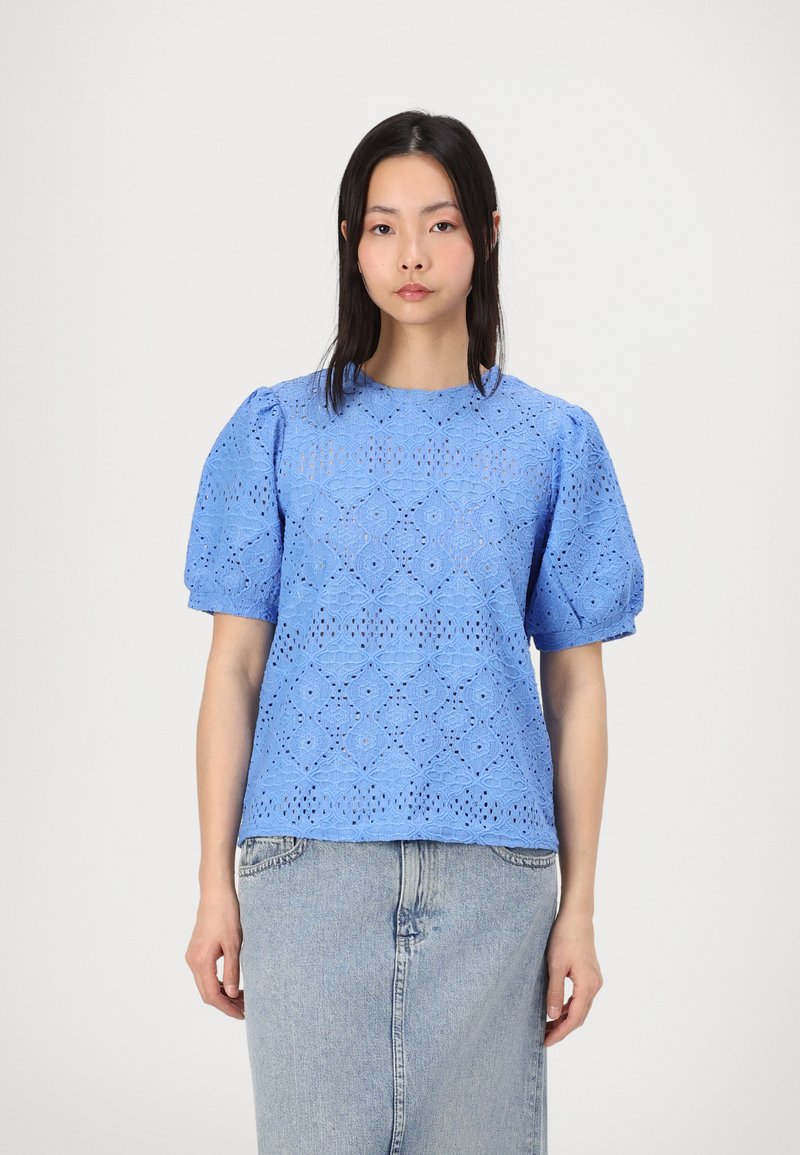 Blouse en coton bleu clair avec des manches bouffantes, ornée d'un motif de dentelle à trous floraux et d'un col rond, portée sur une jupe en denim.