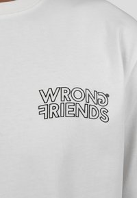 Bílá mikina se nápisem "WRONG FRIENDS" tučným černým písmem. Design zahrnuje kulaté písmo s odlišnými styly písmen.