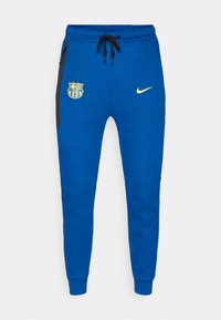 Calças de jogging em algodão azul, apresentando um bolso com fecho éclair preto, o logo do FC Barcelona e o swoosh da Nike em amarelo. Punhos ajustados nos tornozelos.