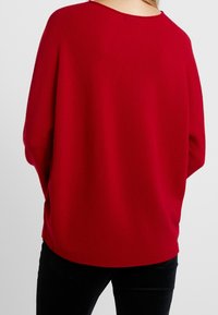 Pull rouge en maille avec une coupe décontractée, un col rond et des épaules tombantes. Le tissu présente une texture lisse et un motif minimaliste.