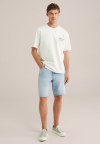 Valkoinen puuvillapaita, jossa on "EXPLORE" -graafinen printti, yhdistetty vaaleansinisiiin denim-shortsit. Mallilla on päällään vaaleanharmaat lenkkarit. Neutraali tausta.