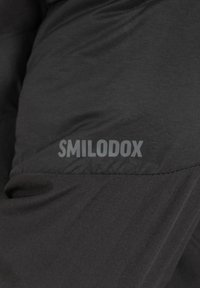 Veste de sport noire avec une texture lisse et mate, portant le logo "SMILODOX" imprimé en gris clair sur la manche supérieure gauche.