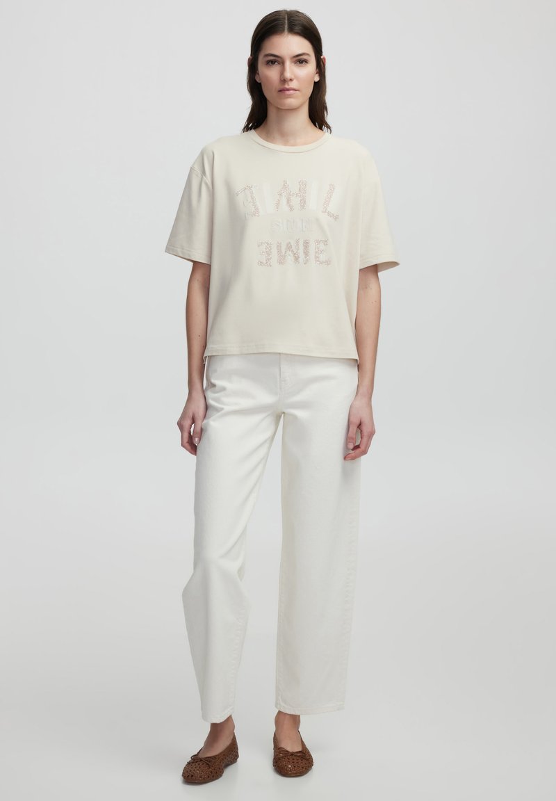 T-shirt cropped beige con lettering testurizzato, abbinata a pantaloni bianchi a vita alta e scarpe slip-on in rafia marrone. Finitura in tessuto liscia.