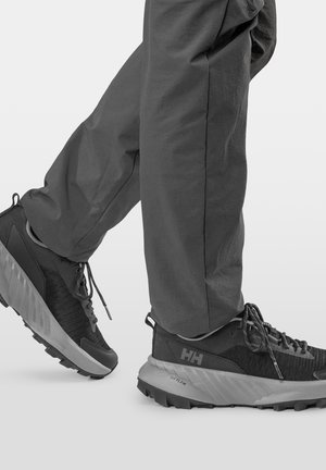 Czarno-szare buty do wędrówek Helly Hansen z teksturowaną podeszwą, noszone z ciemnymi spodniami na jednolitym tle.