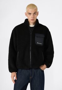 Chaqueta de forro polar negra con cierre frontal, cuello alto y bolsillo en el pecho. Textura suave con el logotipo de Champion y material de bolsillo en contraste.