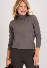 Grijze coltrui van een zachte breigaren, met lange mouwen en geribde manchetten, gecombineerd met donkere broek en een armband.