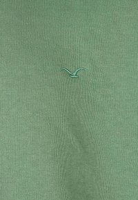 Light green T-shirt with a ribbed texture and an embroidered bird logo on the left side. Simple design, soft fabric.