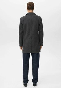 Manteau en laine gris avec un col classique, une coupe ajustée, une fente unique et un design droit. Porté avec un pantalon bleu foncé et des chaussures assorties.
