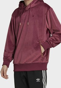 Tummanpunainen huppari, jossa on sileä pinta, kullanväriset metalliosat, kiristysnauhoilla varustettu huppu ja vetoketjullinen sivutasku. Resorit hihansuissa ja helmassa.