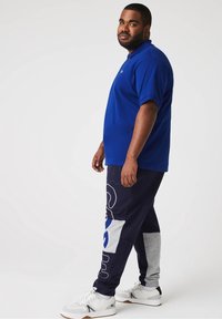 Homme debout de profil portant un polo bleu royal, un pantalon colorblock marine et gris, et des baskets blanches sur fond uni clair.