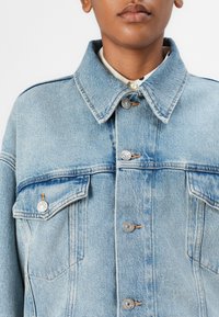 Lichtblauwe denim jas met een kraag, zilveren knopen, twee voorzakken en een lichte vervaging voor textuur.