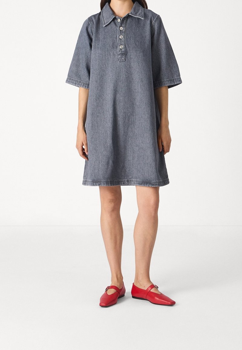 Personne portant une robe en jean gris, ample, longueur genoux, avec des boutons et des chaussures plates rouges sur un fond blanc uni.