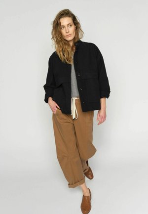 Laineliste juustega naine kannab musta nööpidega jopet, halli särk, pruune lahtisi pükse nööriga vöökohal ning pruune punutud kingi, seisab ühevärvilise tausta ees.