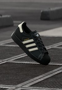 Μαύρο αθλητικό παπούτσι Adidas Superstar με λευκές ρίγες και χρυσή ετικέτα «Superstar» που αιωρείται πάνω από υφή μεταλλικών σχαρών σε εξωτερικό χώρο.