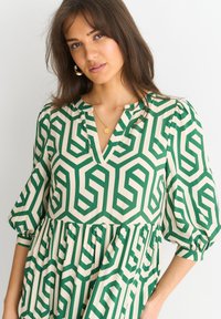 Green and cream patterned dress with geometric designs, featuring a V-neck, puff sleeves, and gathered waistline. Gold necklace visible.