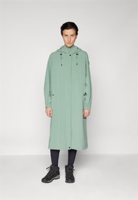 Long manteau vert menthe avec une capuche, doté de cordons de serrage noirs et de boutons, porté sur des leggings foncés et des bottes à lacets noires.