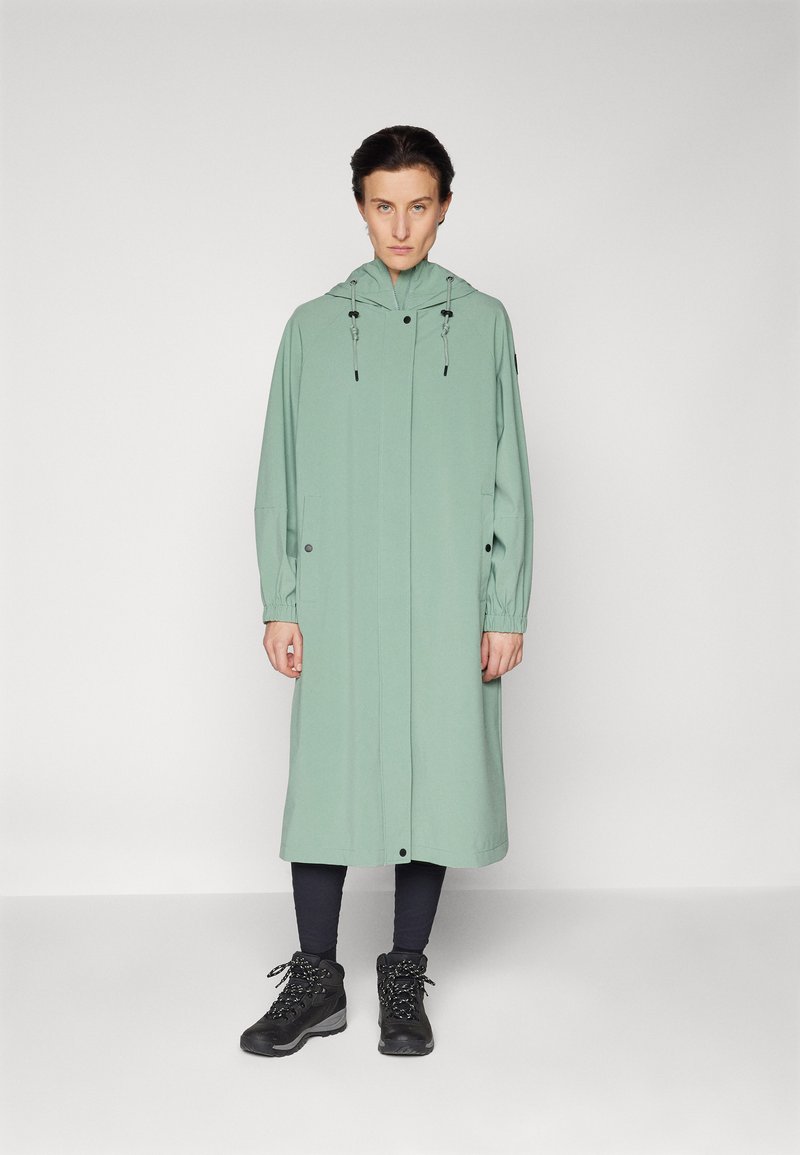 Long manteau vert menthe avec une capuche, doté de cordons de serrage noirs et de boutons, porté sur des leggings foncés et des bottes à lacets noires.