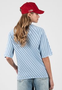 Femme aux longs cheveux ondulés portant une casquette rouge, une chemise à manches courtes bleu clair à motifs et un jean bleu délavé, vue de dos.