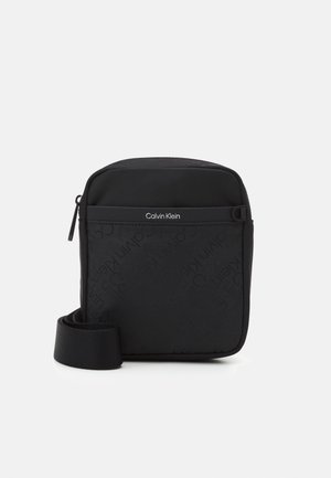 Černá crossbody taška s texturovaným povrchem, na kterém se opakuje vzor loga. Zapínání na zip a nastavitelný popruh. Minimalistický design.