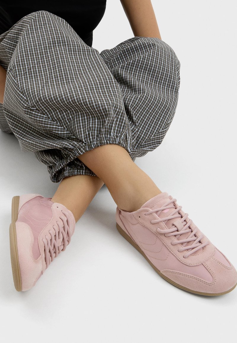 Personne portant un pantalon élastique gris à carreaux avec poignets et des baskets lacées rose clair, assise les jambes croisées sur une surface blanche.