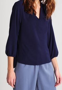 Blouse bleu marine avec col en V, manches ballon trois-quarts, texture lisse et coupe décontractée. Associée à un pantalon large bleu clair.