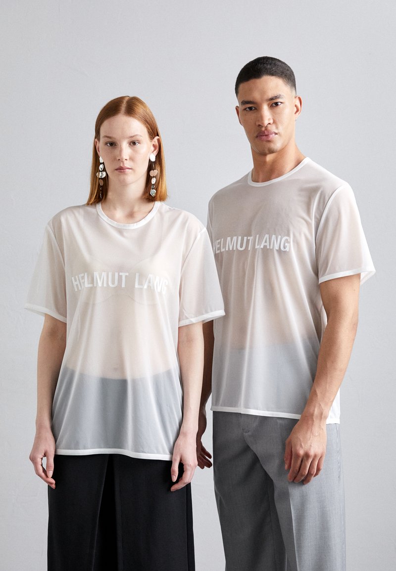 Deux personnes portent des t-shirts Helmut Lang blancs et transparents avec une coupe décontractée. Les t-shirts affichent un logo subtil et contrastent avec des couches inférieures grises.