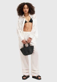 Witte oversized shirt, zwarte bikini top, witte broek met accenten, geweven zwarte handtas, zwarte sandalen; model met krullend haar staat zelfverzekerd.