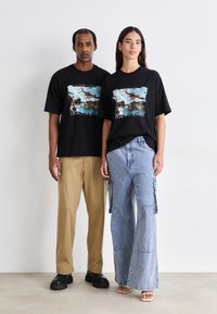 Two models wear black graphic t-shirts featuring a "Legacy of Icarus" print. One pairs it with beige wide-leg pants, the other with blue flared cargo jeans.