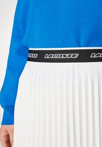 Jupe plissée blanche avec une taille noire arborant le logo "LACOSTE". Le haut est en tissu tricot bleu uni.