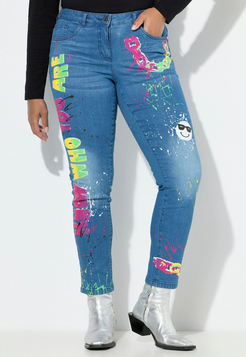 Pantalones de mezclilla azul con estampados gráficos coloridos, que incluyen texto y salpicaduras en rosa, verde y negro, con un ajuste ceñido y un bolsillo trasero.