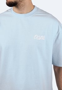 Světle modré bavlněné tričko s kulatým výstřihem, krátkými rukávy a bílým vyšitým logem "CRYSTAL" na levé straně hrudi. Měkká textura.