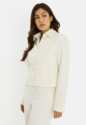 Mujer de cabello largo y oscuro, vestida con un blazer crema abotonado con bolsillos delanteros y pantalones a juego, mirando hacia un lado sobre un fondo liso.