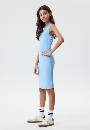 Pige med langt mørkt hår iført en ærmeløs lyseblå kjole til knæene, hvide sokker med sorte striber og hvide sneakers, stående i profil.