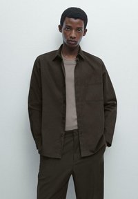 Dark brown button-up shirt with a chest pocket, worn over a light brown fitted shirt. The outfit features a contemporary, minimalist style.