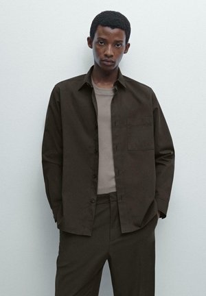 Dark brown button-up shirt with a chest pocket, worn over a light brown fitted shirt. The outfit features a contemporary, minimalist style.