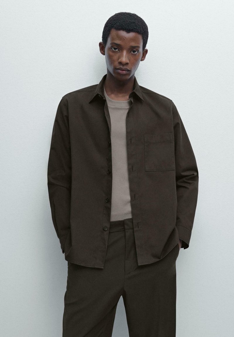 Dark brown button-up shirt with a chest pocket, worn over a light brown fitted shirt. The outfit features a contemporary, minimalist style.