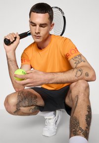 Vyras oranžine marškiniais ir juodomis šortais sėdi ant kelių, laiko teniso kamuoliuką ir raketę, rodo tatuiruotes ant rankų ir kojų.