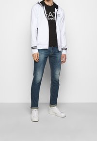 Felpa bianca con zip e rifiniture nere, t-shirt grafica nera, jeans blu e sneaker bianche. Outfit casual con colori e materiali a contrasto.