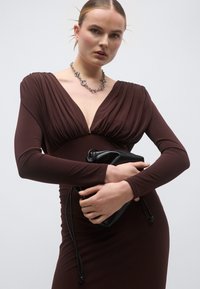 Καφέ φόρεμα μακρυμάνικο με βαθύ V-λαιμόκοψη, ύφασμα που πλαισιώνει το λαιμό. Μαύρη τσάντα στο χέρι, ασημένιο κολιέ με αλυσίδα προσθέτει λεπτομέρεια.