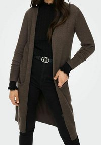 Brązowy swetrowy kardigan na drutach z długimi rękawami, otwarty z przodu, z dwoma kieszeniami i miękką fakturą, zestawiony z czarnym topem i dżinsami.