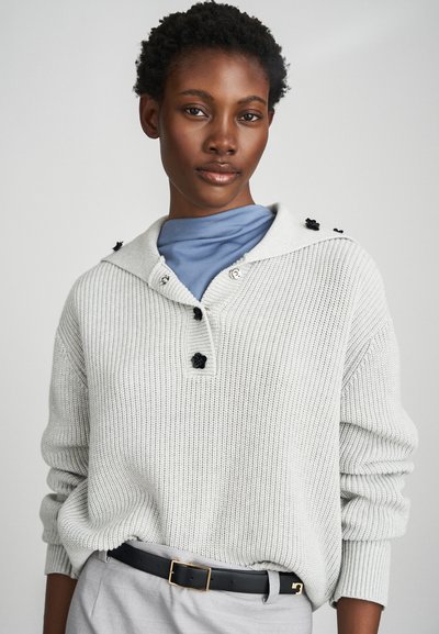 Svjetlosiva pletena džemper s šalom, crni cvjetni gumbi i dugi rukavi. Nosi se preko plave majice i uparen je s crnim remenom.