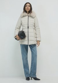 Piumino beige chiaro con colletto in pelliccia sintetica, vita cinturata e chiusura con zip. Abbinato a jeans chiari e scarpe con tacco nere.